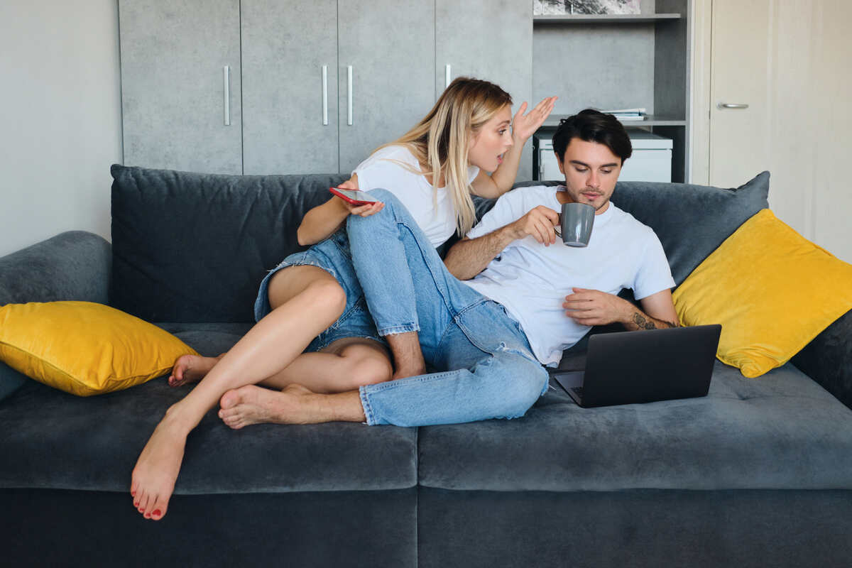 angry blond woman with cellphone in hand emotionally talking to brunette man with cup of coffee and laptop