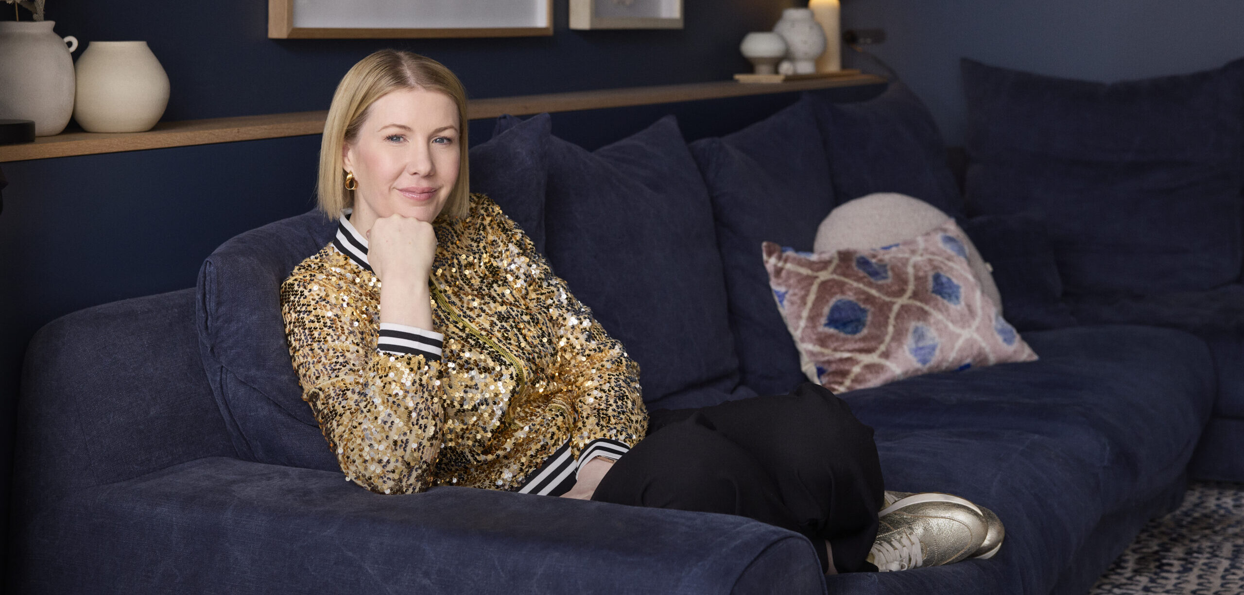 Meg sits on blue couch wearing a gold jacket, looking toward the camera with a smile.