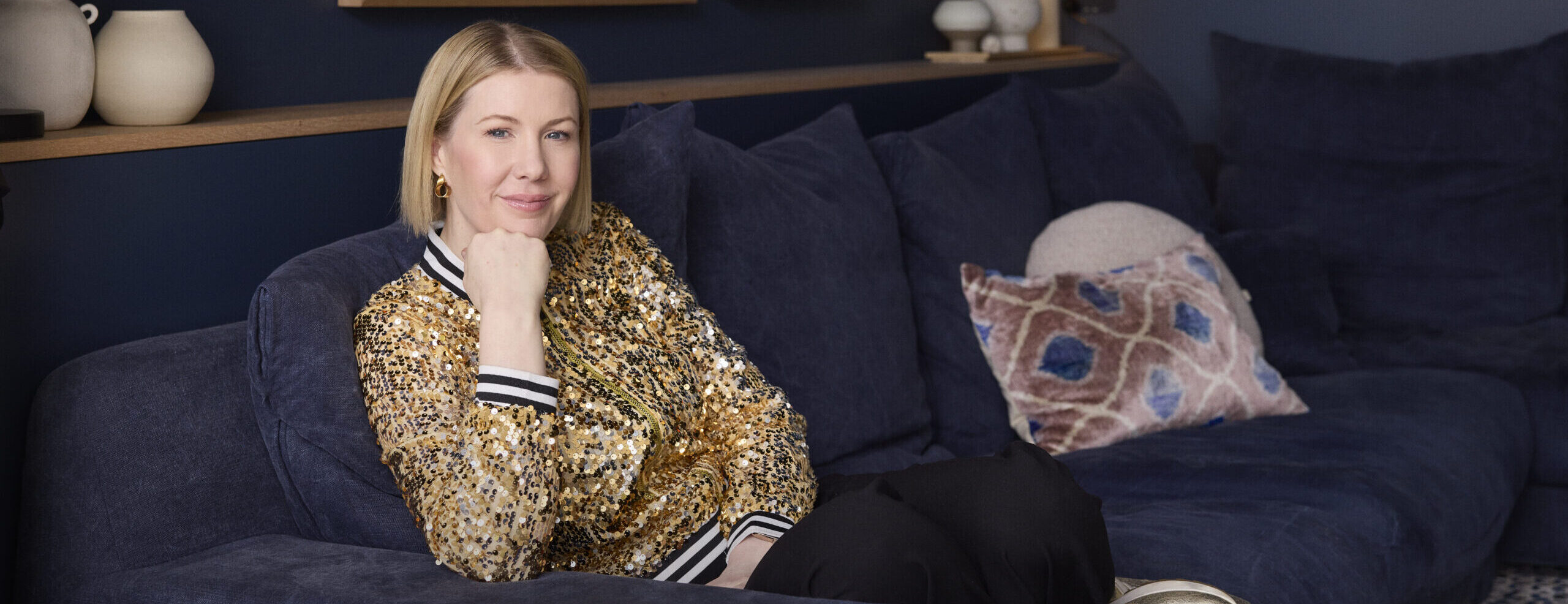 Meg sits on blue couch wearing a gold jacket, looking toward the camera with a smile.